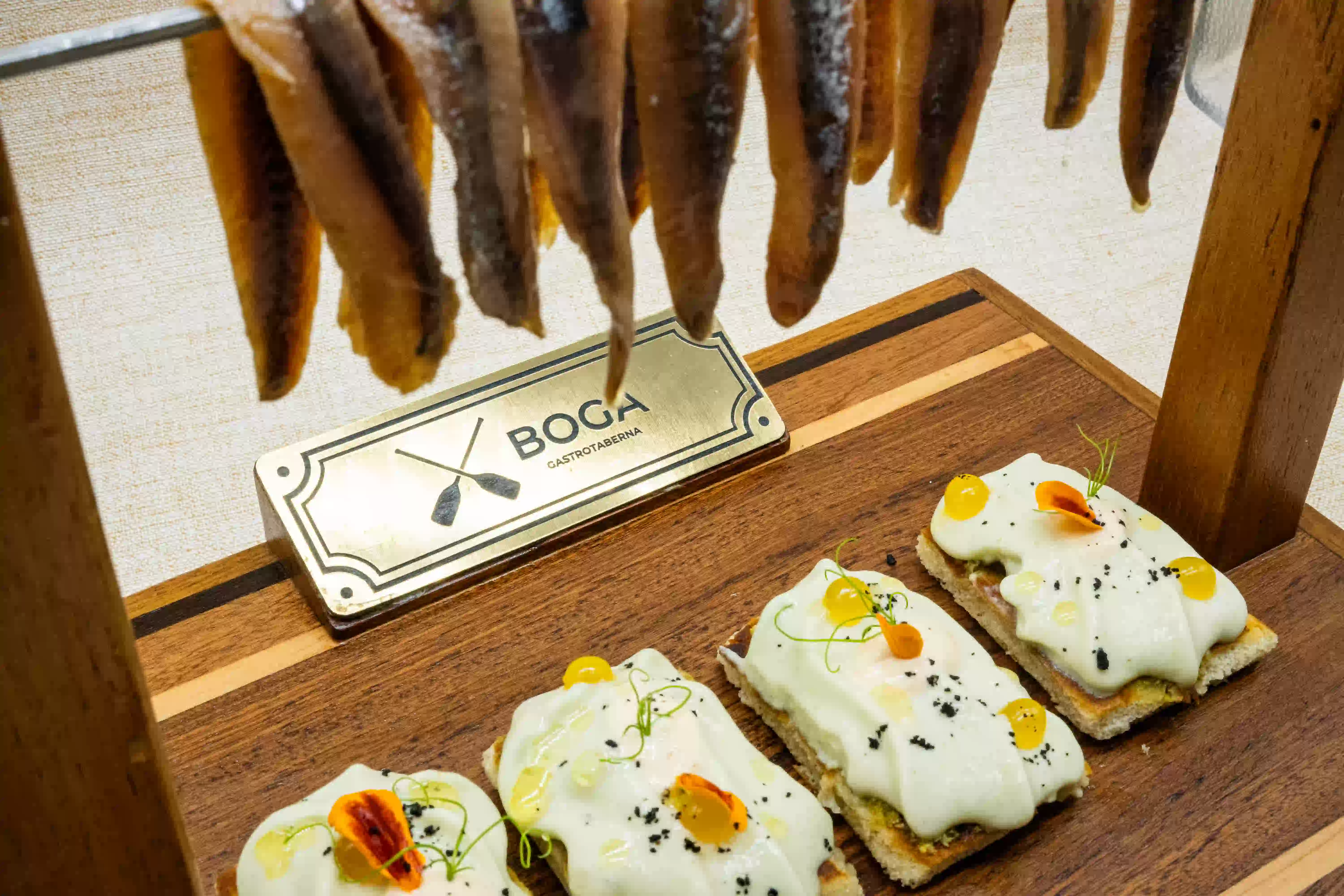 Anchoas colgadas sobre una tabla de madera con cuatro tostas de anchoa, crema y huevas, junto a la placa de Boga